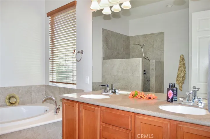 Primary bath with easy-access, walk-in, tiled shower with extra-tall dual shower heads; matching tile surround on soaking tub, dual sinks, Corian-style countertop, wood-look blinds, and tile flooring.