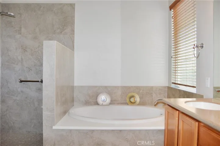 Primary bath with easy-access, walk-in, tiled shower with extra-tall dual shower heads; matching tile surround on soaking tub, dual sinks, Corian-style countertop, wood-look blinds, and tile flooring.