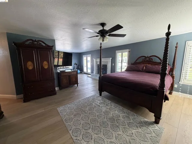 Bedroom featuring light wood finished floors, a textured ceiling, ceiling fan, a fireplace with raised hearth, and access to outside