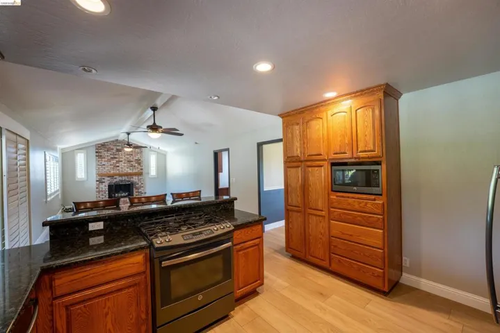 Kitchen featuring stainless steel appliances, a brick fireplace, wood finish cabinetry, dark stone counters, and beam ceiling