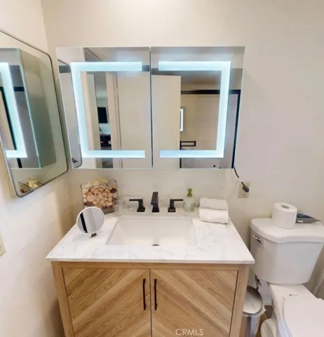 Spa-like bathroom featuring an updated vanity with a modern sink, marble-look countertop, and warm wood cabinetry, paired with a Hollywood-style LED double mirror with built-in interior mirrors. A zero-threshold walk-in shower with built-in bench and detachable shower head, along with a wall-mounted towel warmer, completes this thoughtfully designed space.