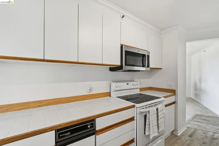 Two tone kitchen featuring white range with electric stovetop, crown molding, tile counters, stainless steel microwave, and light wood finished floors