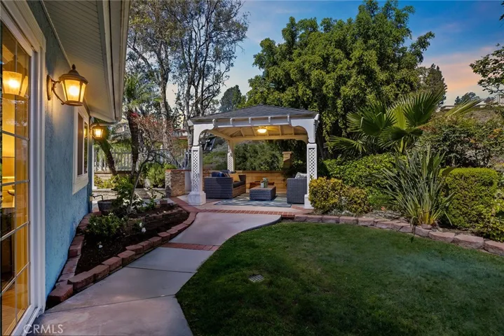 As you come through the picket fence, this is your backdrop—the backyard comes to life in the evening with soft lighting, a peaceful setting, and the gazebo ready for a quiet night or time with friends and family.