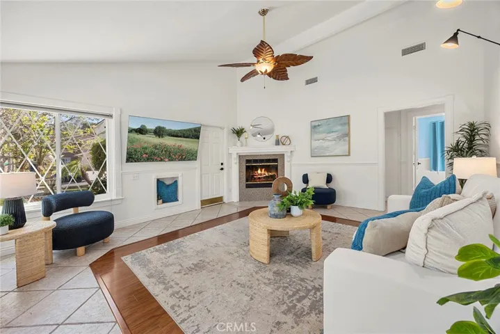 Bright, inviting living room with vaulted ceilings, a ceiling fan, and a cozy fireplace. The front door features decorative glass that brings in natural light, and the room looks out to the front and side yard with a nice sense of privacy. Fireplace has been digitally enhanced for presentation.