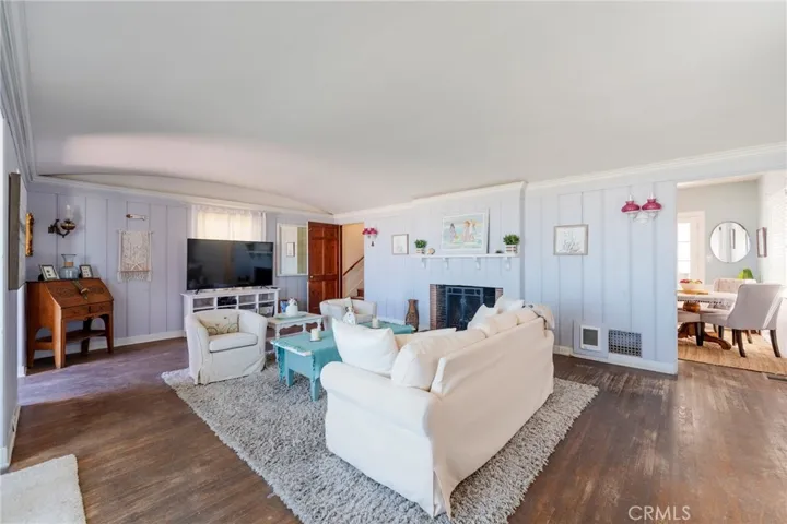 Bright, coastal-inspired living room with open layout, wood floors, and a cozy fireplace. Light-filled space flows seamlessly to dining area, featuring soft tones and inviting seating—perfect for relaxing or entertaining.