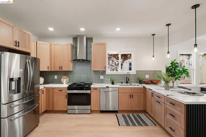 Kitchen featuring stainless steel appliances, a peninsula, light brown cabinetry, wall chimney range hood, and hanging light fixtures