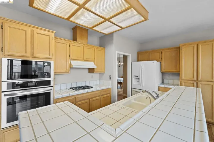 Kitchen featuring tile countertops, white appliances, light wood finish cabinets, and a kitchen island with sink