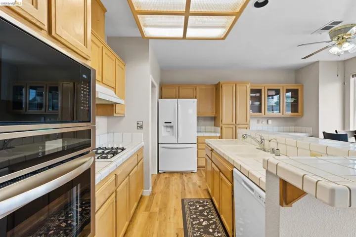 Kitchen with tile counters, white appliances, light wood-style flooring, light wood finish cabinetry, and a ceiling fan