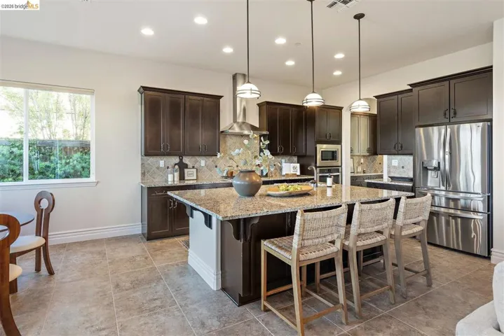 Kitchen featuring dark wood finish cabinetry, stainless steel appliances, a kitchen breakfast bar, a kitchen island with sink, and light stone counters