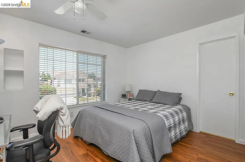Room featuring wood-finish flooring, a white ceiling fan with integrated lighting, a spacious window with horizontal blinds, and built-in shelving