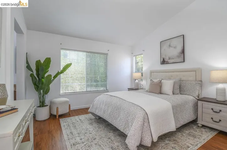 Vaulted ceilings define this bright bedroom, featuring wood-finish flooring, a large window with horizontal blinds, and an upholstered headboard