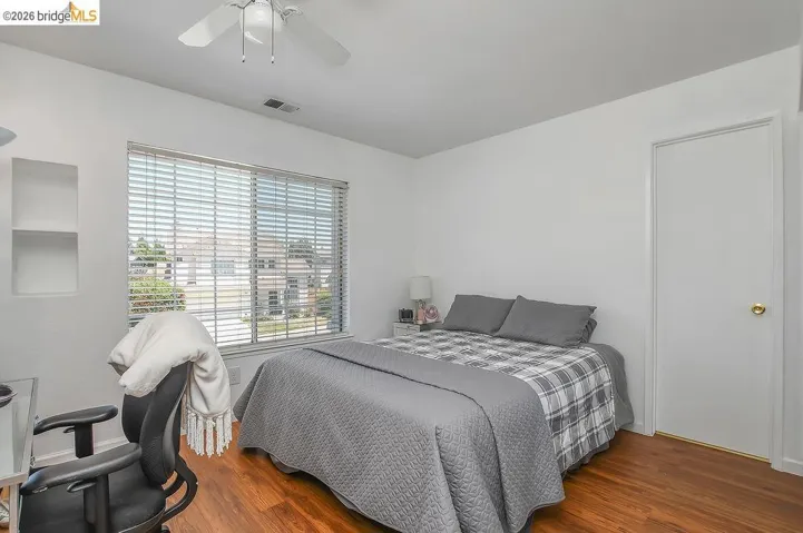 Room featuring wood-finish flooring, a white ceiling fan with integrated lighting, a spacious window with horizontal blinds, and built-in shelving