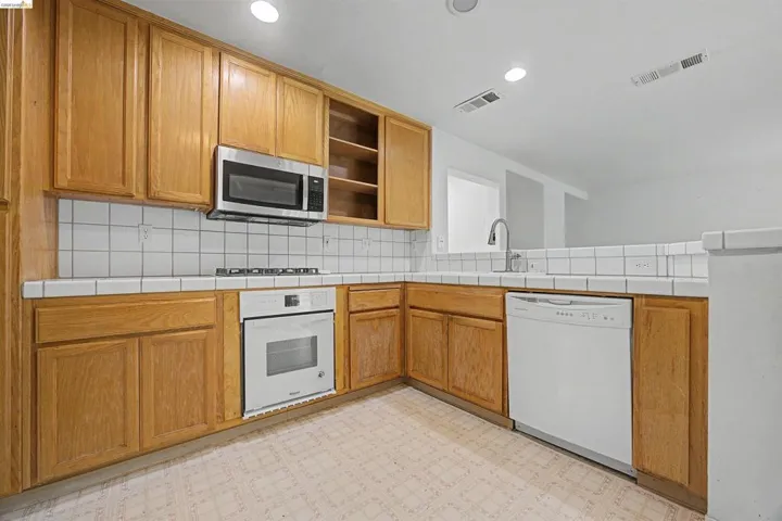 Kitchen with tile counters, open shelves, backsplash, white appliances, and light floors