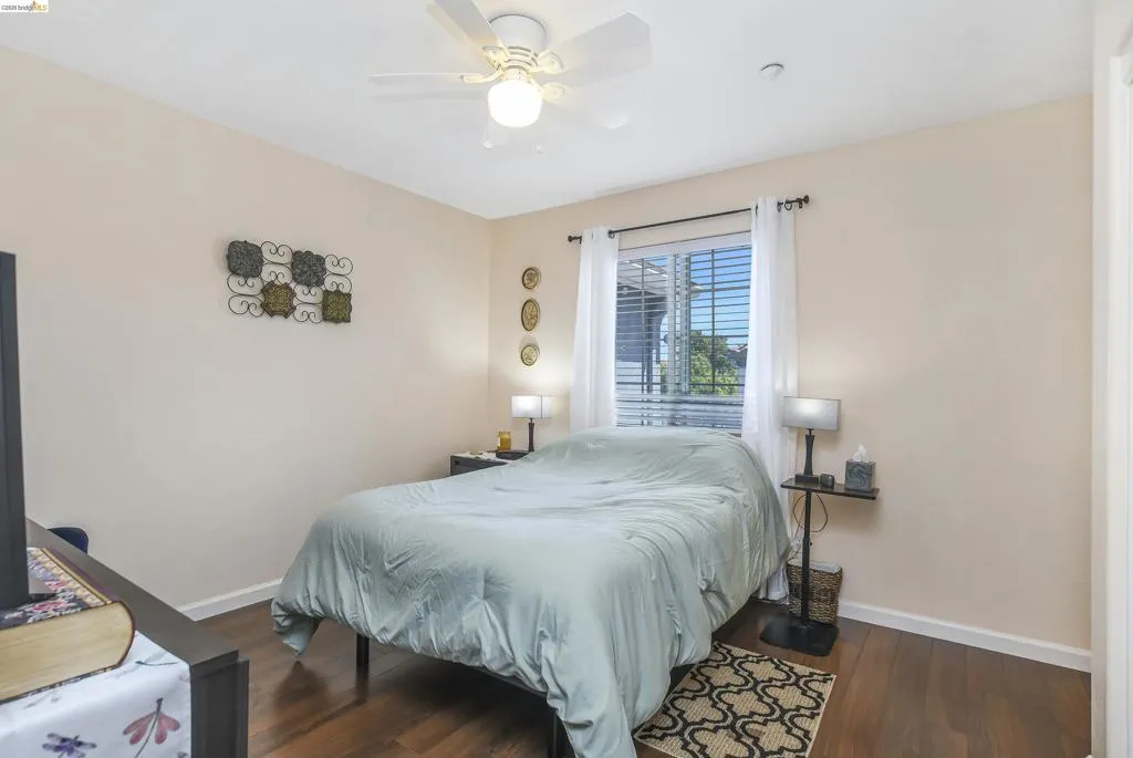 Bedroom featuring wood-finish flooring, a ceiling fan with integrated lighting, and a window with horizontal blinds and sheer white curtains