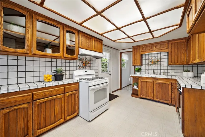 Sunlit Kitchen with an eat-in dining area, tile countertops, gas range, vent hood, dishwasher, and abundant natural wood cabinetry.