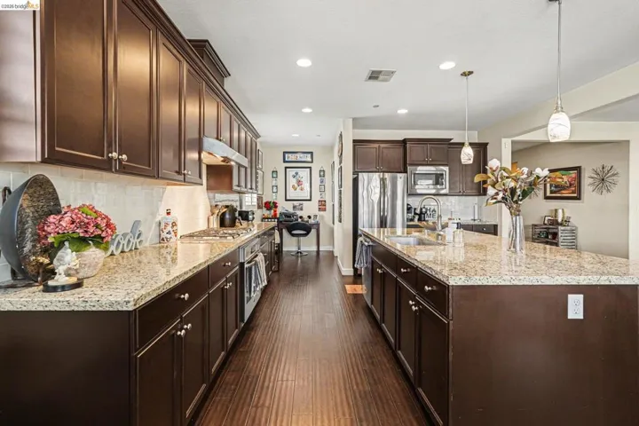 Kitchen with dark wood finish cabinets, dark wood-type flooring, decorative light fixtures, light stone counters, and an island with sink