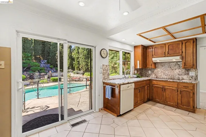 Kitchen featuring wood finish cabinets, dishwasher, granite counters and backsplash, light tile patterned floors, ornamental molding, dual pane slider and window and electric cooktop