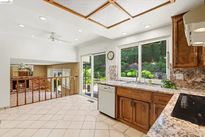 Kitchen with wood finish cabinets, a ceiling fan, range hood, dishwasher, black electric stovetop, granite counter tops and backsplash, tile flooring, dual pane window and slider.