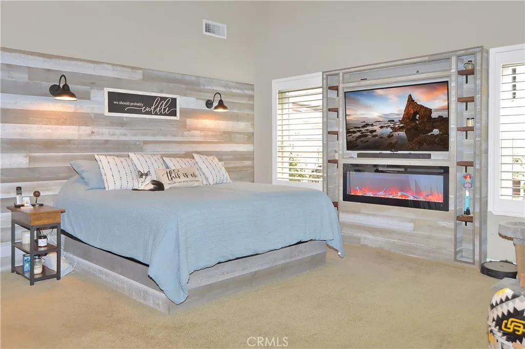 The spacious main bedroom suite boasts more custom built-ins with another heated fireplace and display shelves surrounding the TV (which is included and on a swivel mount for viewing from bed, too), a matching lighted headboard (included), brick accent wall with cabinet and shelving, dramatic cathedral ceilings, louvered shutters, a fantastic fan, and a barn door leading to the en suite bath. (TV image added virtually)