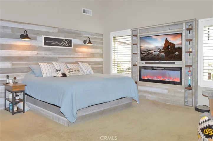 The spacious main bedroom suite boasts more custom built-ins with another heated fireplace and display shelves surrounding the TV (included), a matching lighted headboard (included), brick accent wall with cabinet and shelving, dramatic cathedral ceilings, louvered shutters, a fantastic fan, and a barn door leading to the en suite bath. (TV image added virtually)