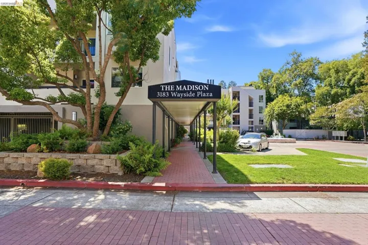 Building exterior featuring a covered walkway with brick-finish flooring, a "THE MADISON" sign, and established landscaping with mature trees