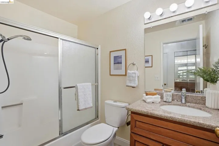 Bathroom featuring a built-in tub with a sliding glass enclosure, a vanity with a granite-style countertop and integrated sink, wood-finish cabinetry, and a wall-mounted mirror with overhead bar lighting