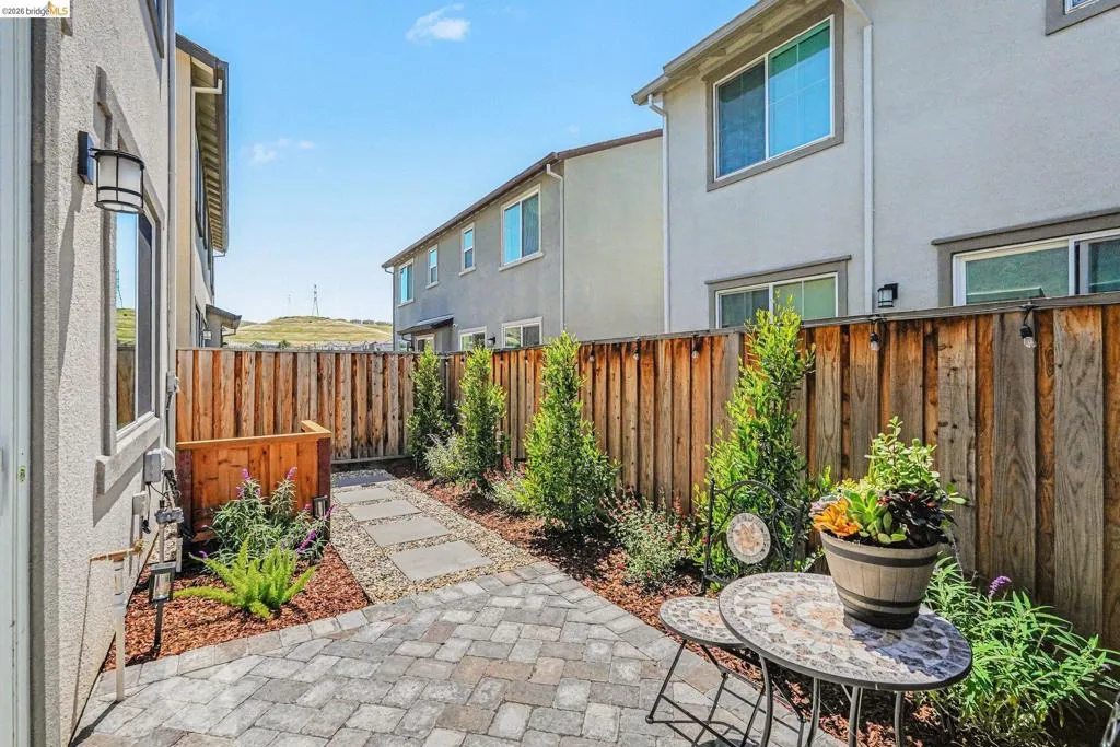 Paver patio area with a stone walkway, bordered by a wood fence and landscaped with mature shrubs and trees