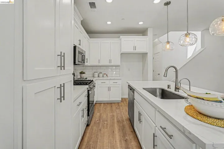 Bright kitchen featuring white shaker cabinetry, stainless steel appliances, wood-finish flooring, recessed lighting, and an island with an undermount sink and pendant lighting