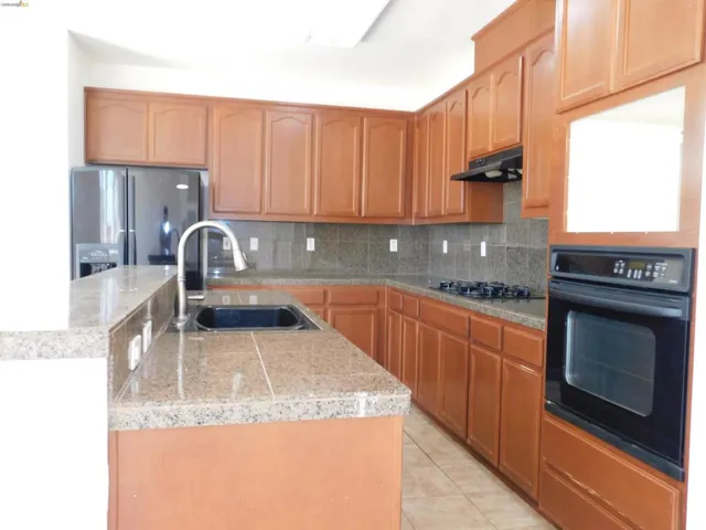 Kitchen featuring black appliances, decorative backsplash, light tile patterned floors, a center island with sink, and wood finish cabinets