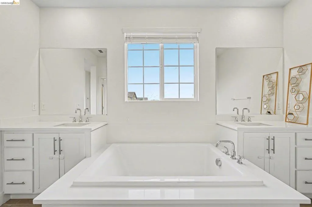 White soaking tub with deck-mounted fixtures, flanked by dual vanities featuring white cabinetry and light-colored countertops
