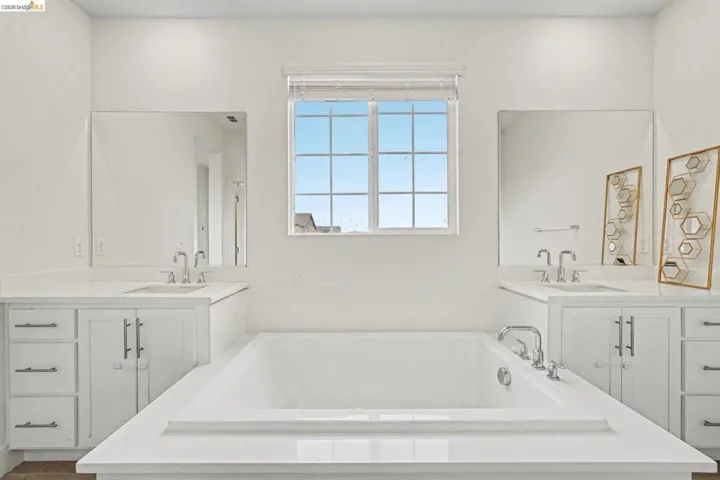 White soaking tub with deck-mounted fixtures, flanked by dual vanities featuring white cabinetry and light-colored countertops