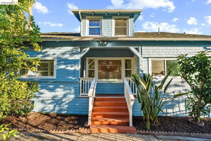 Light blue shingle siding exterior featuring a covered entry porch with white railings, a dormer window, and mature landscaping
