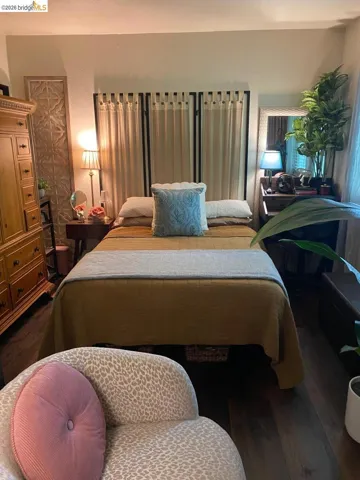 Bedroom featuring dark wood-finish flooring, light-colored walls, and a large window with privacy drapes