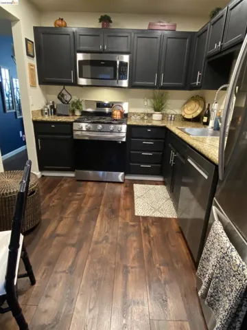 Kitchen featuring dark wood-finish flooring, black cabinetry, granite-style countertops, stainless steel appliances, and a stainless steel sink with a gooseneck faucet