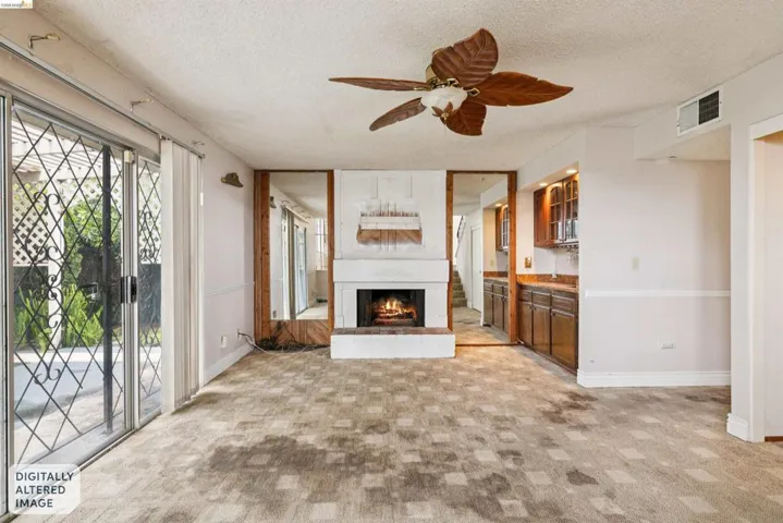 Living area featuring a fireplace with white mantel and brick hearth, patterned carpet flooring, and a ceiling fan with leaf-shaped blades
