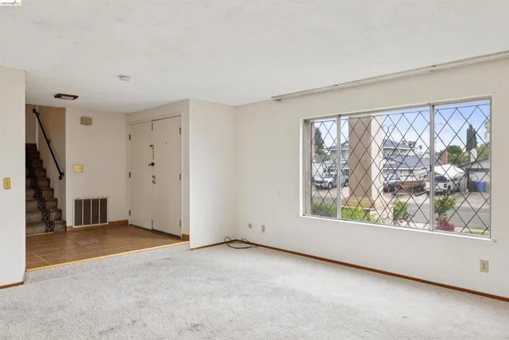 Living area featuring a large window with decorative diamond-patterned security bars, light-toned carpet, and baseboard trim
