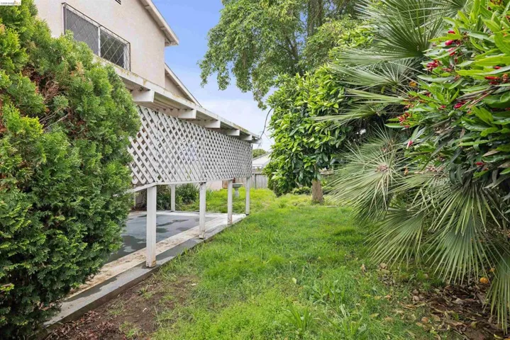 Exterior side yard featuring a covered patio with lattice detailing, mature landscaping, and a grassy area