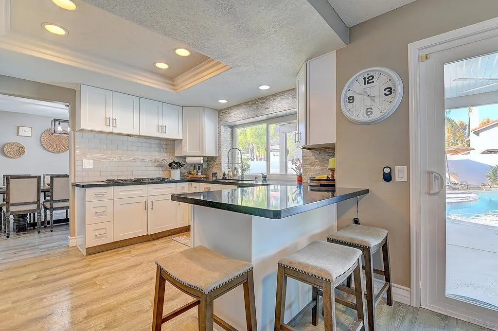 Open, bright, and fully equipped. White shaker cabinetry, granite peninsula with bar seating, tray ceiling with recessed lighting, and a direct door to the backyard and pool — the kitchen that keeps the party going.