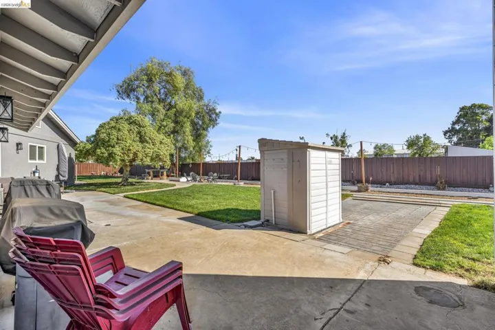 Expansive concrete patio area with a covered section, bordered by a large grassy lawn, a storage shed, and a privacy fence