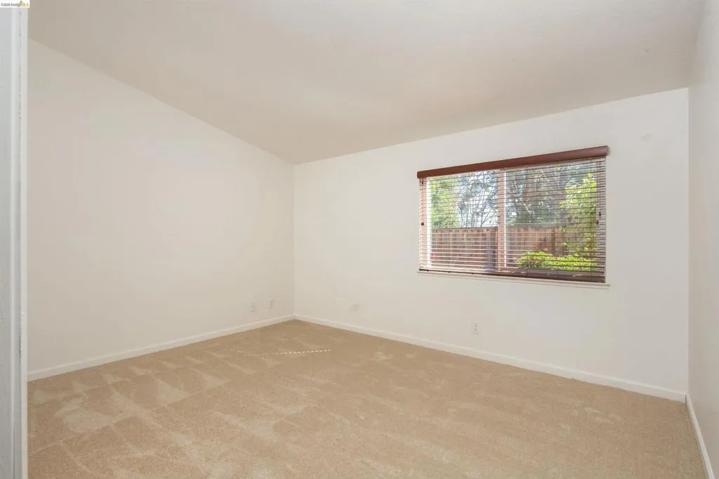 Spacious room featuring neutral carpeting, white walls, a large window with horizontal blinds, and a vaulted ceiling