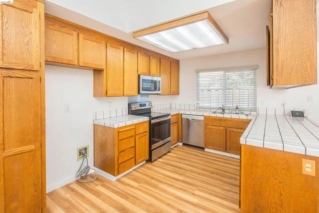 Kitchen featuring light wood-finish cabinetry, stainless steel appliances, a tile countertop with matching backsplash, and wood-finish flooring