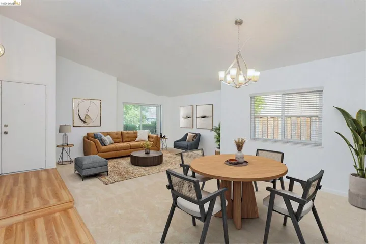 Open-concept interior featuring vaulted ceilings, light-tone carpeting, wood-finish flooring at the entry, and a contemporary chandelier