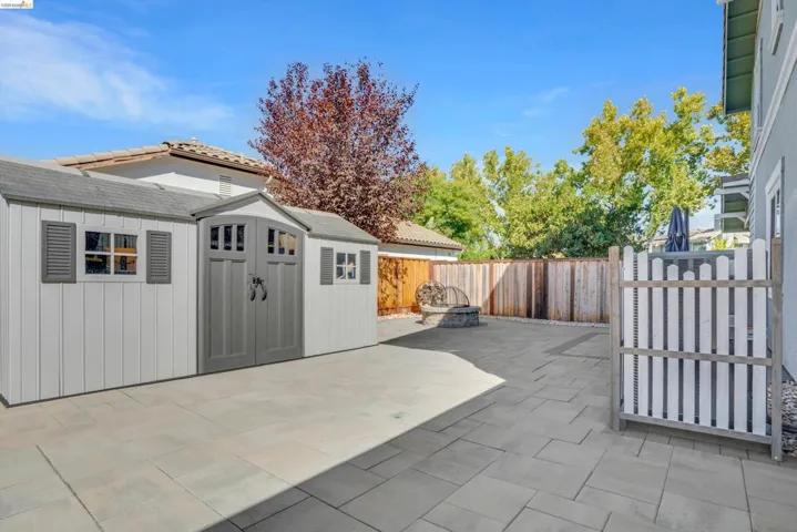 Spacious backyard featuring a large shed with decorative shutters and double doors, expansive paver patio, wood fencing, and a built-in fire pit