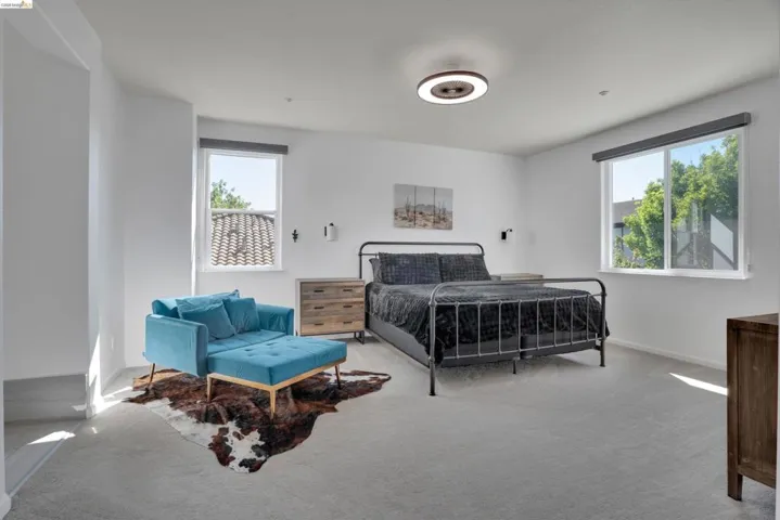 Spacious bedroom featuring light gray carpeting, crisp white walls, two large windows, a modern ceiling light fixture, and two wall-mounted sconces