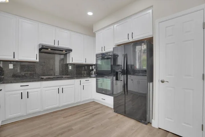 Kitchen featuring wood-finish flooring, white cabinetry with dark hardware, a dark built-in double oven, a dark side-by-side refrigerator, and dark countertops with a matching backsplash