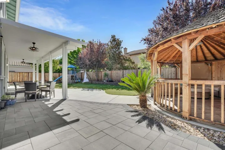Expansive backyard featuring a covered patio with ceiling fans, a spacious paved area, a manicured lawn, a wooden gazebo, and mature landscaping