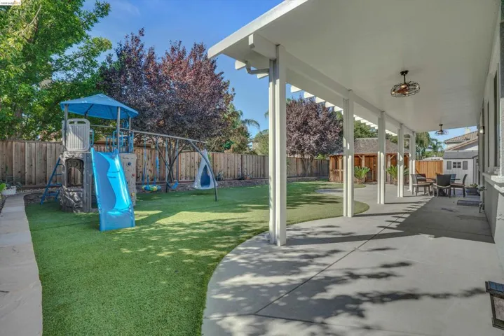 Expansive backyard featuring a covered patio, artificial turf, wood fencing, mature trees, and a concrete pathway