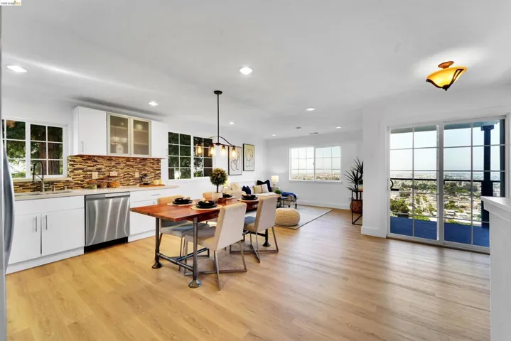 Open-concept living space featuring wood-finish flooring, a contemporary kitchen with white cabinetry and stainless steel appliances, a stacked stone backsplash, and expansive sliding glass doors opening to a cityscape