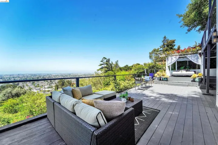 Expansive outdoor deck featuring wood-finish flooring, a glass railing overlooking the distant cityscape, and a built-in pergola structure with white privacy drapes