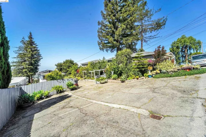 Expansive driveway with sloped grade, featuring mature trees, diverse plantings, a picket fence, and distant architectural elements